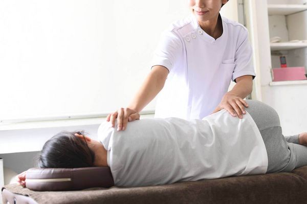 Ein Therapeut in weißer Kleidung führt eine Rückenmassage bei einer Person in seitlicher Position aus.