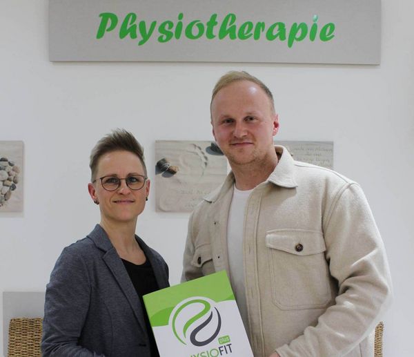 Zwei Personen stehen lächelnd in einer Physiotherapiepraxis in Helmstedt und halten eine Mappe mit dem Logo von PhysioFit.